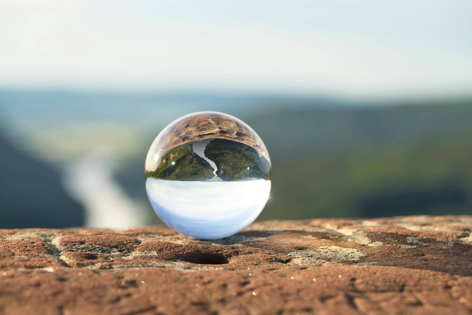 A clear glass sphere rests on a rocky surface, reflecting an upside-down view of a river winding through green hills. The background is softly blurred, drawing focus to the crisp, detailed landscape within the sphere.