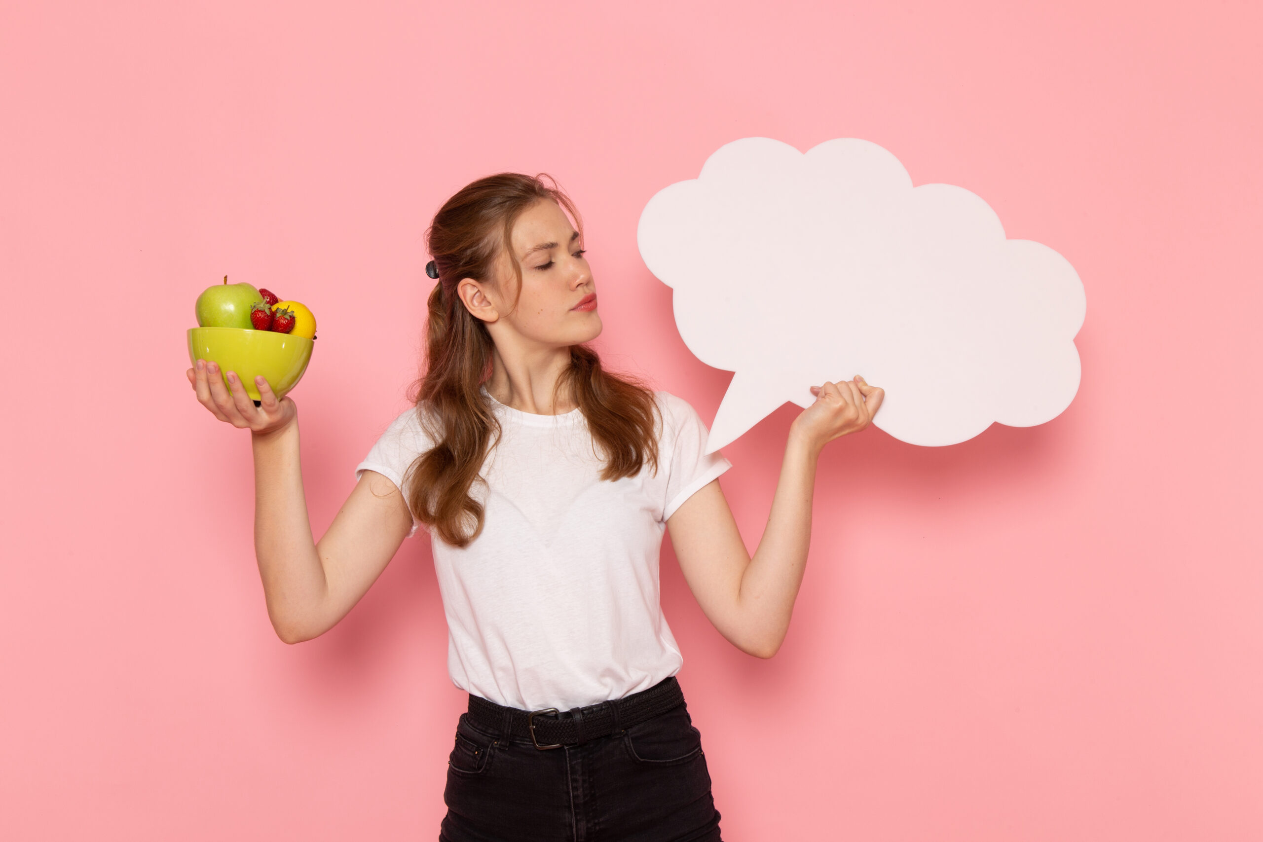 Front view of young female in white t-shirt holding a bowl of fruit and a dialogue sign.